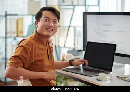 Portrait of a cheerful senior developer working at a desk with a computer and screens displaying programming code. This technology-themed image highlights software development, IT professions, and corporate workplaces. Stock Photo