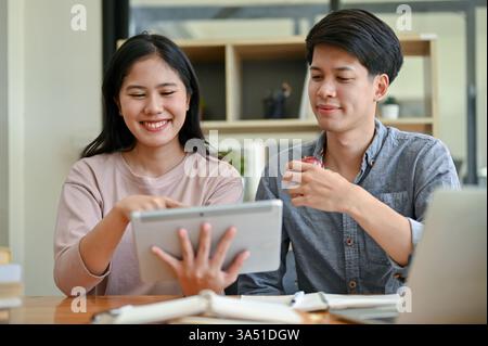 Two cheerful Asian college students look at a tablet together, discussing ideas for a group project in a campus cafe setting. The image highlights collaboration and study in diverse student life. Ideal for education, teamwork and campus life content. Stock Photo