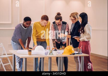 Busy diverse group of architects and engineers collaborate over blueprints on a table in a modern office. Emphasizes teamwork, design, and planning in architecture and construction contexts. Includes a strong copy space for headlines and captions. Stock Photo