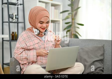 Smiling Asian Muslim woman wearing a hijab relaxes at home. She eats cookies while watching a movie on a laptop, with headphones resting on her neck on the sofa. Ideal for home lifestyle, remote work and streaming content visuals. Stock Photo