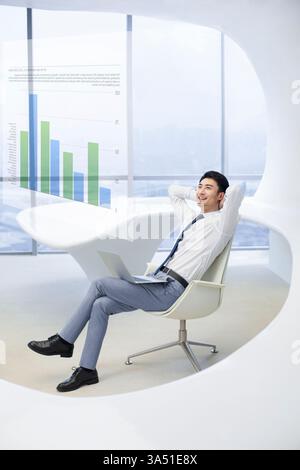 Modern Chinese businessman in his 20s using a touch screen in a bright office. The scene emphasizes business technology with data visuals like bar graphs and financial figures, suggesting forecasting and analysis. A clean, professional setting with a laptop and windowed backdrop conveys confidence and innovation. Stock Photo