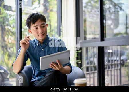 Thai millennial man sits in a cafe, thinking about his savings plan while using a tablet. The casual urban coffee shop setting highlights remote working and personal planning. This image is ideal for articles on budgeting, fintech, and freelance lifestyles. Stock Photo