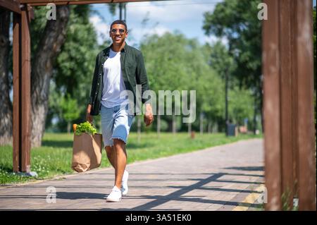 Smiling African American man wearing sunglasses holding a bag of food while strolling in a sunny park. A cheerful outdoor lifestyle image with natural light, greenery and a relaxed mood, ideal for wellness, food or leisure campaigns. Suitable for editorial or commercial use showcasing positive vibes and outdoor activity. Stock Photo