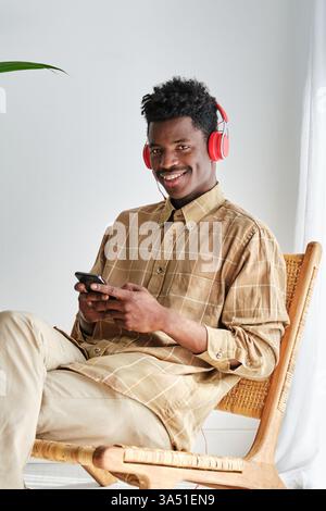 Cheerful African American man wearing a modern headset and looking at the camera while sitting with legs crossed on a wicker chair. A contemporary lifestyle portrait perfect for customer service, online support or informal work-from-home scenes. Ideal for tech and social media visuals. Stock Photo