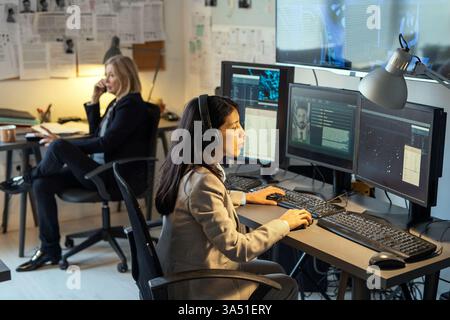 Serious female intelligence officer sits at a desk in a modern office, focused on computer monitors and data. This high tech scene suits stories about cyber security, crime investigation, and government agencies. A professional stock photo for law enforcement, intelligence, and data processing topics. Stock Photo