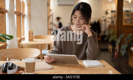 Focused young Asian woman uses a digital tablet in a cozy cafe, studying online and working remotely. This lifestyle scene suits content about remote work, online learning, and cafe productivity in the city. Ideal for marketing or editorial visuals featuring freelancers, students, and modern workspace on the go. Stock Photo
