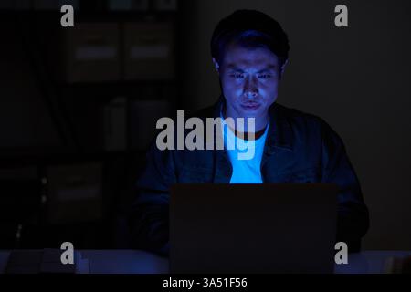 Portrait of a serious Asian man working on a laptop in a dark room at night. The moody tech scene suits cybersecurity, coding, and late-night work themes. Stock Photo