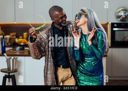 Smiling Black man looks at his Asian wife wearing oversized sunglasses and sticking out his tongue in a playful kitchen scene at home. Lighthearted couple lifestyle image perfect for love, humor, and domestic life content. Stock Photo