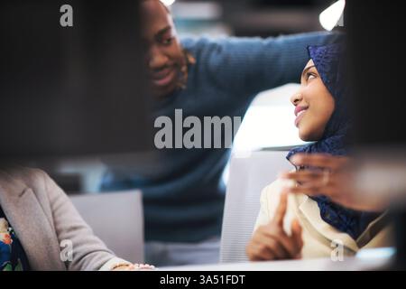 Smiling Black Muslim woman wearing a hijab participates in a meeting with coworkers at a table in the office. The diverse team collaborates in a professional setting with laptops and notes. Suitable for diversity, teamwork, and corporate culture visuals. Stock Photo