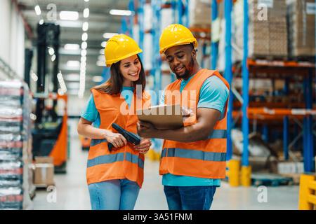 Black male worker wearing hard hat and safety vest holding clipboard and managing inventory with caucasian female coworker while standing in warehouse Stock Photo