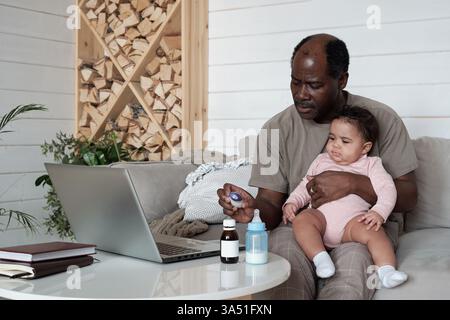 Horizontal shot of a Black man sitting on a sofa at home caring for his baby with a common cold. A tender fatherhood moment focused on family health and attentive parenting. Stock Photo