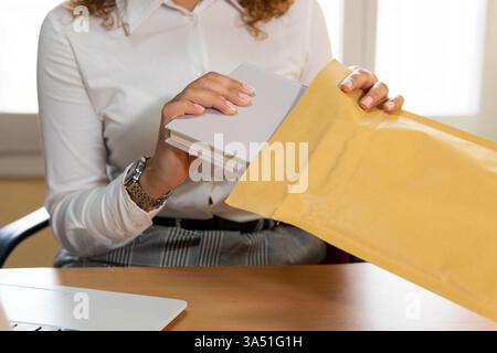 Faceless Hispanic female entrepreneur places an agenda into an envelope at a desk with a laptop in an office. This professional scene with copy space is ideal for business communications and startup visuals. Stock Photo