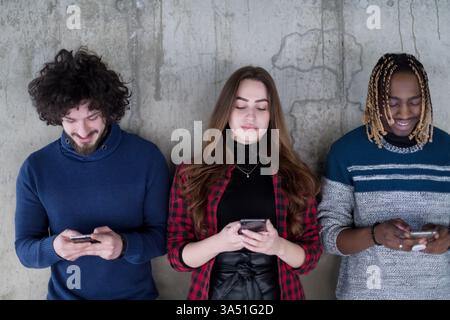 Diverse group of young professionals casually dressed, using smartphones in front of a concrete wall at a modern startup office. Great for teamwork, collaboration and technology themes in a contemporary workplace. Stock Photo