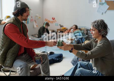Mature woman taking cup of tea from hands of young male refugee while both sitting in front of camera in crisis camp Stock Photo