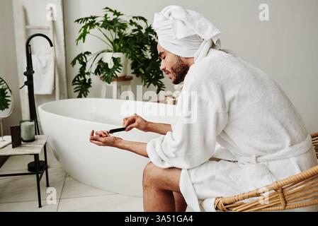 Young man in a bathrobe performs a self manicure with a nail file in a bathroom. Personal care and wellness imagery for grooming and self-care topics. Stock Photo