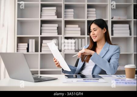 Professional and confident millennial Asian businesswoman reading and reviewing business reports at her desk in a private office. She sits at a tidy desk, examining paperwork and company reports, conveying corporate, finance, and professional themes. This image suits business, workplace, and office communication contexts, with a focus on Asian professionals. Stock Photo