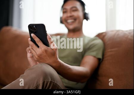 A happy millennial Asian man relaxes on a brown leather sofa at home, using his smartphone. The casual setting and modern lifestyle capture everyday tech use and leisure. Ideal for technology, mobile, and home living visuals. Stock Photo