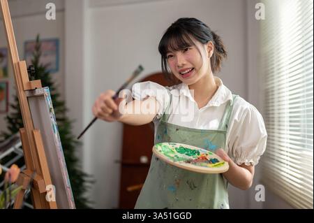 Creative young Asian woman artist in a home studio holding a paint brush and color palette. She stands by an easel, ready to create in a light-filled space. Stock Photo