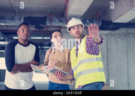 Architect or engineer uses a tablet to show house plans to a young, multiethnic couple at a construction site. This image suits architectural design, home building projects and client consultations. Stock Photo