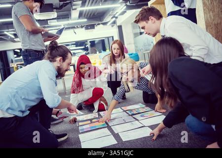 Diverse group of business professionals brainstorm and present ideas on the office floor. They collaborate in a modern workspace, highlighting teamwork, startup culture, and cross-cultural communication. Stock Photo
