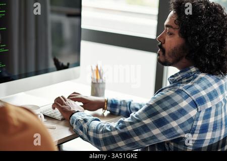 A focused Indian programmer works at a desk in a modern office, coding on his computer. The scene conveys software development, IT and technology in a professional workspace. Stock Photo