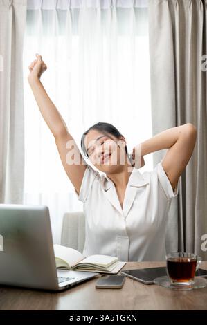 Carefree young Asian woman at her desk, stretching her arms after finishing a task in a modern office. This relaxed moment highlights wellbeing and work-life balance for corporate and remote-work campaigns. Great for business lifestyle, employee wellbeing and productivity imagery. Stock Photo