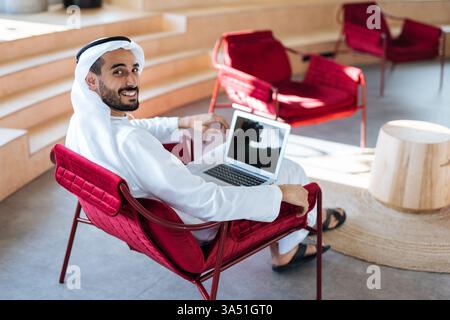 Smiling Arab man sits on a couch in a bright office and chats on a laptop during the day. This professional, remote-work scene is ideal for stories about Middle Eastern business culture and modern corporate communication. It conveys a confident, connected vibe suitable for UAE and Gulf business content. Stock Photo