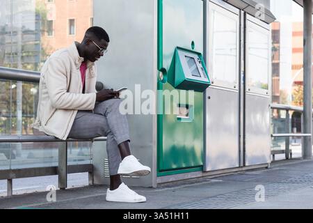 African American man waiting at a tram station while using a mobile phone. The urban transport setting suits travel, commuting, and everyday lifestyle themes. Perfect for infrastructure, public transport, or workplace commuting contexts. Stock Photo