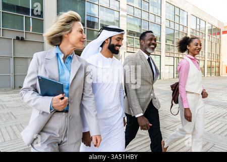 A diverse group of business professionals bonds outdoors in the city, led by an Emirati businessman. The scene highlights international collaboration and corporate teamwork in a modern urban setting. They wear smart business attire and appear engaged in discussion. Stock Photo