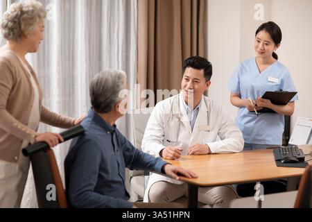 Chinese male doctor talks with a senior male patient, accompanied by his wife and a nurse in a clinic. A candid healthcare moment suitable for medical services, patient care, and family involvement. Stock Photo