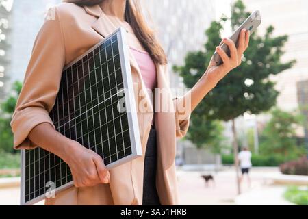 From below view of an unrecognizable female entrepreneur in a blazer standing on a city street with a portable solar panel while texting on a mobile phone. The image emphasizes entrepreneurship, technology, and renewable energy. Ideal for green business and mobile communications themes. Stock Photo