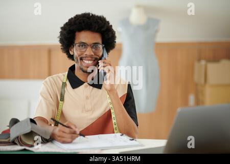 Portrait of a happy fashion designer in a studio, sketching a design while talking on the phone with a client. This image captures the creative process in a fashion atelier and suits editorial content on design and small business. Stock Photo