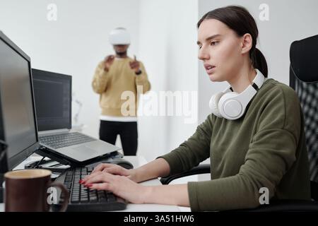 Two colleagues in a tech-focused office. A mixed-race woman wearing headphones works at a computer while a Black male colleague stands nearby wearing a VR headset. Stock Photo