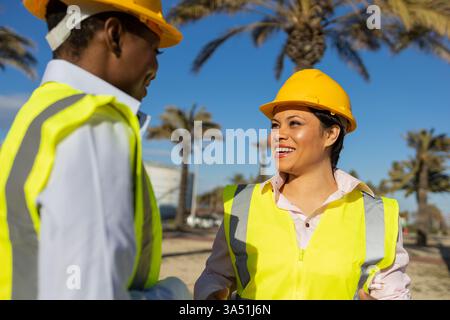 Two women engineers, one Hispanic and one Black, shake hands in front of a construction site on a sunny day, wearing hard hats and safety vests. Highlights diversity in engineering and professional collaboration in a field setting. Ideal for STEM, construction, teamwork and women in industry campaigns. Stock Photo
