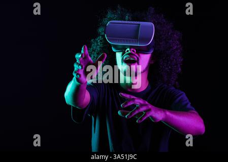 Latino man wearing VR goggles stands with arms raised in a dark room illuminated by neon light. The scene highlights immersive virtual reality and futuristic tech vibes. Perfect for technology, innovation and digital lifestyle campaigns. Stock Photo