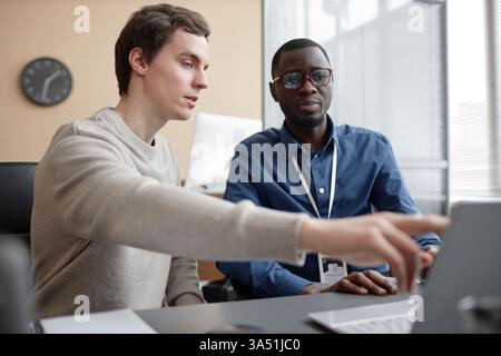 Waist-up shot of a young Caucasian man pointing at a laptop screen while speaking with an African American coworker in a modern office. A diverse, collaborative workplace scene suitable for technology, IT, and team-work campaigns. Stock Photo