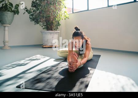 Full-body shot of a focused young Asian woman in athletic wear performing a yoga pose on a mat. A serene studio scene highlighting flexibility, balance, and mindfulness. Stock Photo