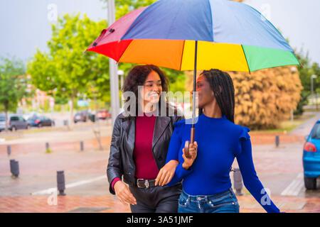 Smiling Black woman walks with a Hispanic friend under a rainbow umbrella on a rainy city street. Colorful urban lifestyle imagery suited to diversity, friendship, and travel or fashion projects. Stock Photo