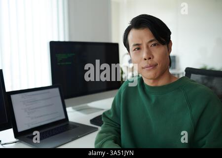 Portrait of a serious Asian software developer seated at his office desk. This professional image emphasizes technology, coding, and the IT workplace, suitable for business and software industry campaigns. Stock Photo