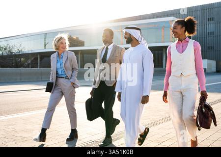 Multiethnic group of business professionals meeting outdoors in the city, led by an Emirati businessman. The scene showcases teamwork, diversity and international collaboration. Bright daylight and urban setting convey professional energy, suitable for corporate communications and cross-cultural marketing. Stock Photo