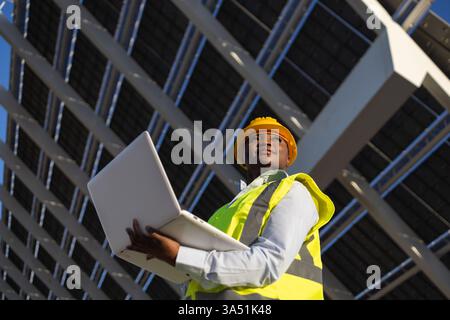 Black female engineer wearing a hard hat and safety vest uses a laptop on a construction site under solar panels. The scene highlights renewable energy, engineering and on site work in a modern industrial setting. Perfect for construction, technology and sustainable development topics. Stock Photo