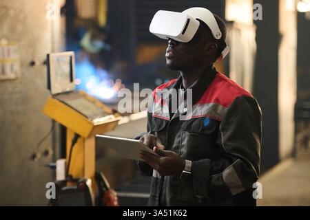 Young African American factory worker wearing a VR headset and using a tablet while operating an industrial machine. The image blends technology with manufacturing in a technical production environment. Perfect for Industry 4.0, worker training and digital manufacturing themes. Stock Photo