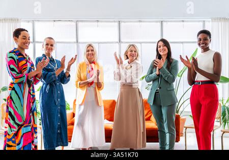 Group of diverse, confident businesswomen meeting in a modern office. A multicultural team collaborates on a brainstorming session, clapping and celebrating ideas. This image is ideal for stories about women in leadership, teamwork, and workplace empowerment. Stock Photo