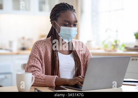 Black professional woman in her 20s works from home on a laptop during COVID-19 quarantine. She wears a protective mask at a kitchen desk in a home office, reflecting remote work during the pandemic. Stock Photo