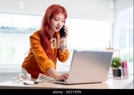 A confident Asian female graphic designer with red curly hair talks on the phone while working on her laptop. She is in a modern office space, focused on design projects and digital work. Stock Photo