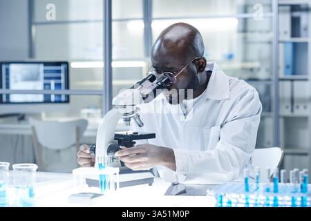 A focused Black male scientist in a lab coat studies samples under a microscope in a modern laboratory. Ideal for topics such as Covid-19 research, genetics, microbiology, and biotechnology in healthcare settings. The image conveys concentration, scientific analysis, and medical innovation. Stock Photo
