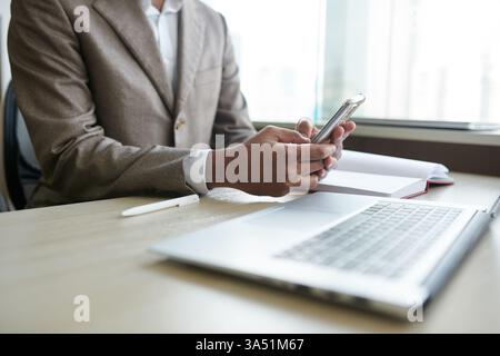 Cropped image of a professional Asian man in a suit checking messages on a smartphone at his desk. A concise, business-ready shot suitable for technology, communication, and workplace themes. Stock Photo