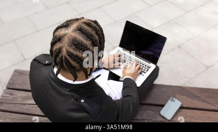 Hispanic man uses a laptop on a park bench during daytime. The scene captures remote work in an urban outdoor setting with portable tech. Ideal for telework, freelancing and digital lifestyle visuals. Stock Photo