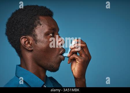 Minimal side-view portrait of a Black man using an asthma inhaler against a blue background with copy space. Clean studio shot ideal for healthcare, medical campaigns, and wellness communications. Stock Photo