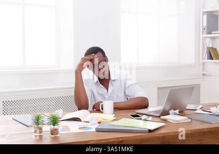 A thoughtful African American employee sits in an office, contemplating a new project with a coffee nearby. The desk and copy space emphasize focus in a contemporary workplace. Great for content about brainstorming, decision making, and professional life. Stock Photo